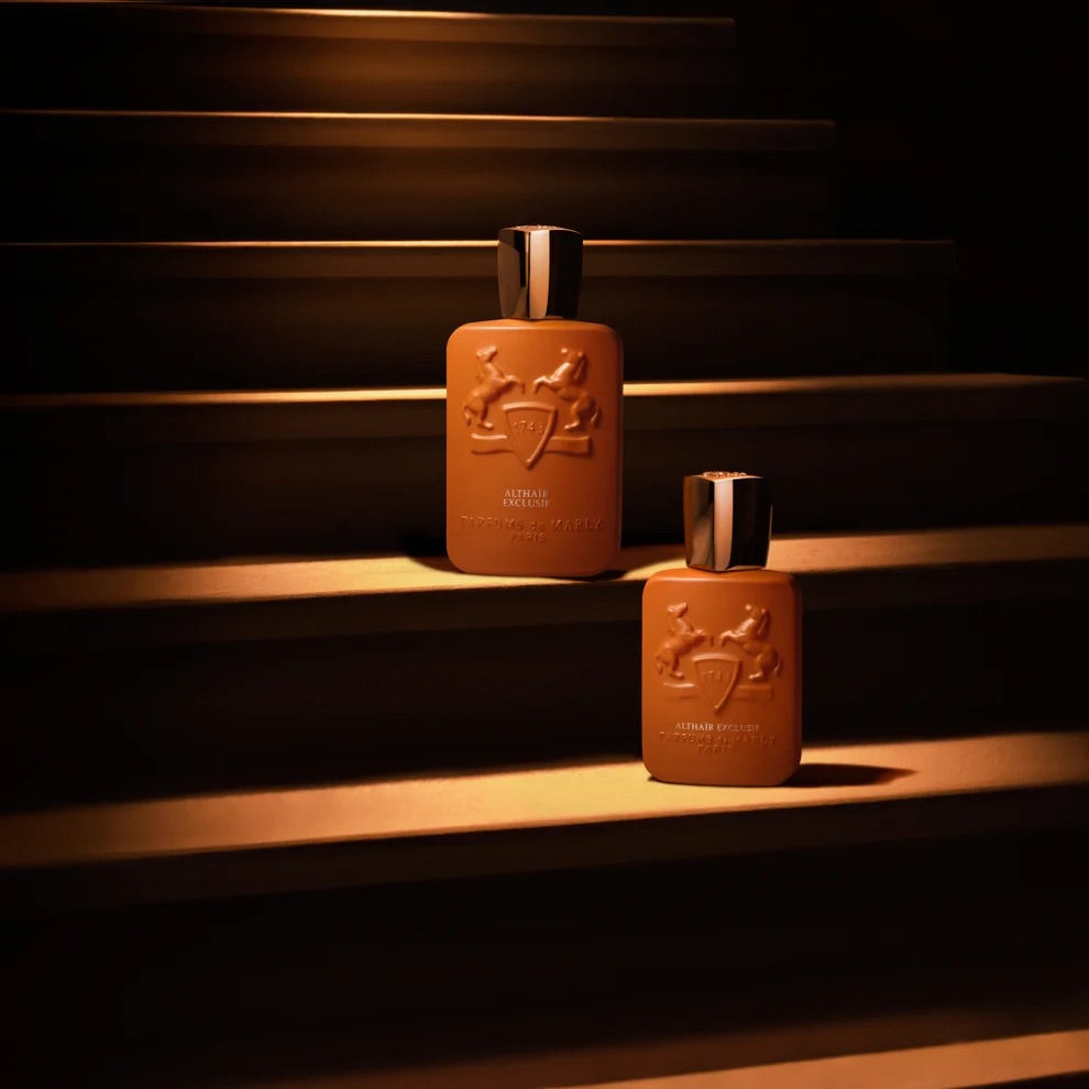 Two amber-brown perfume bottles with metallic silver caps, featuring an embossed lion logo, displayed on dark wooden stairs under dramatic warm lighting.