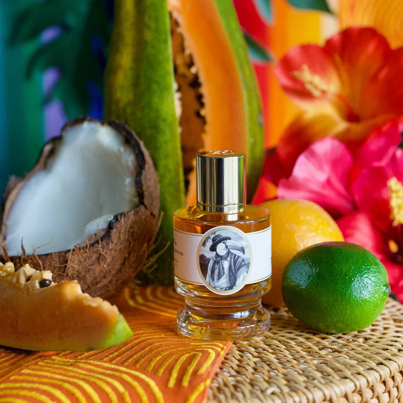 A small clear glass perfume bottle with a metallic cap and a white label featuring a black-and-white portrait, styled among tropical fruits and flowers on a woven mat. A cut coconut, lime, orange, papaya, and vivid flowers fill the background.