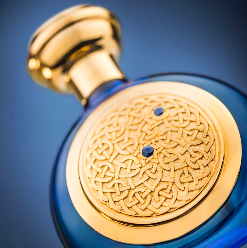 A close-up of an ornate perfume bottle with a deep blue glass body, a polished gold cap, and a large circular gold medallion featuring an intricate knot pattern and two small blue gem accents, set against a dark blue gradient background.
