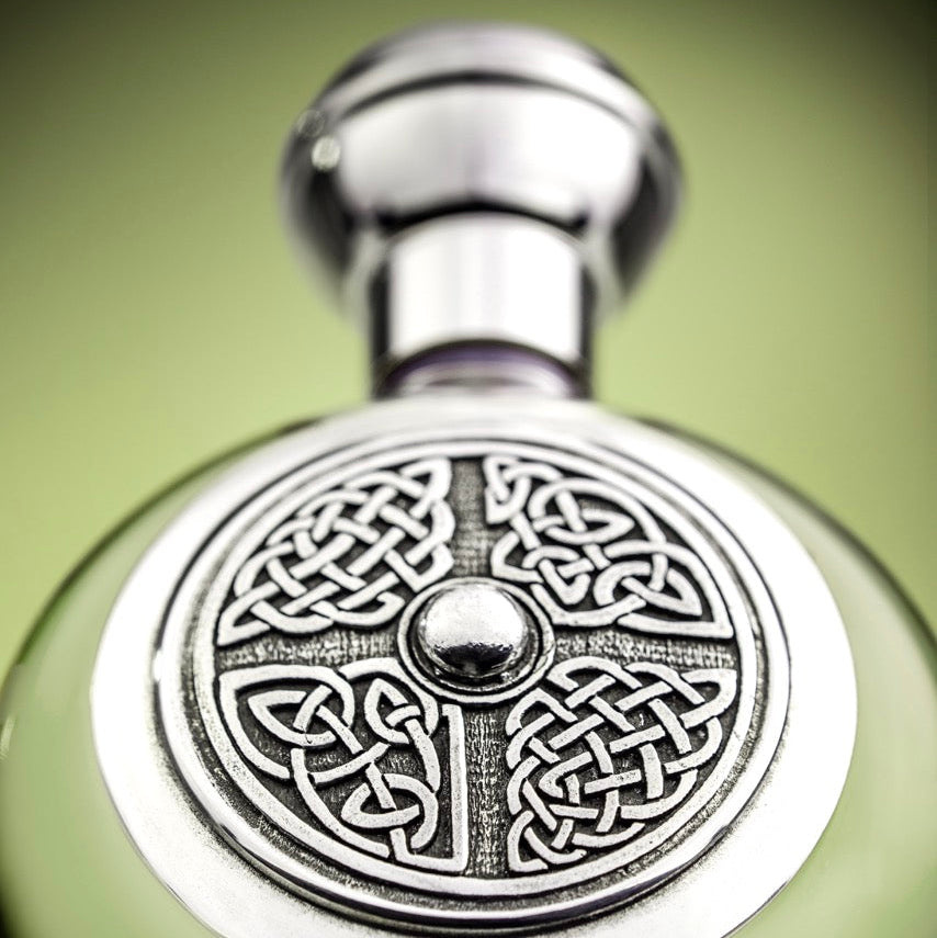 A close-up of a decorative glass bottle with a polished silver-toned cap and a circular Celtic knot pattern on the front, set against a soft green gradient background.