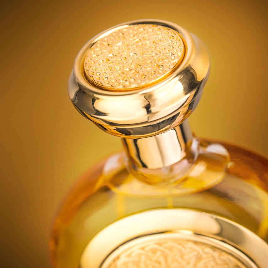 A close-up of a decorative amber glass bottle with a shiny gold-toned stopper and embossed detailing on the bottle body, set against a warm golden-brown gradient background.