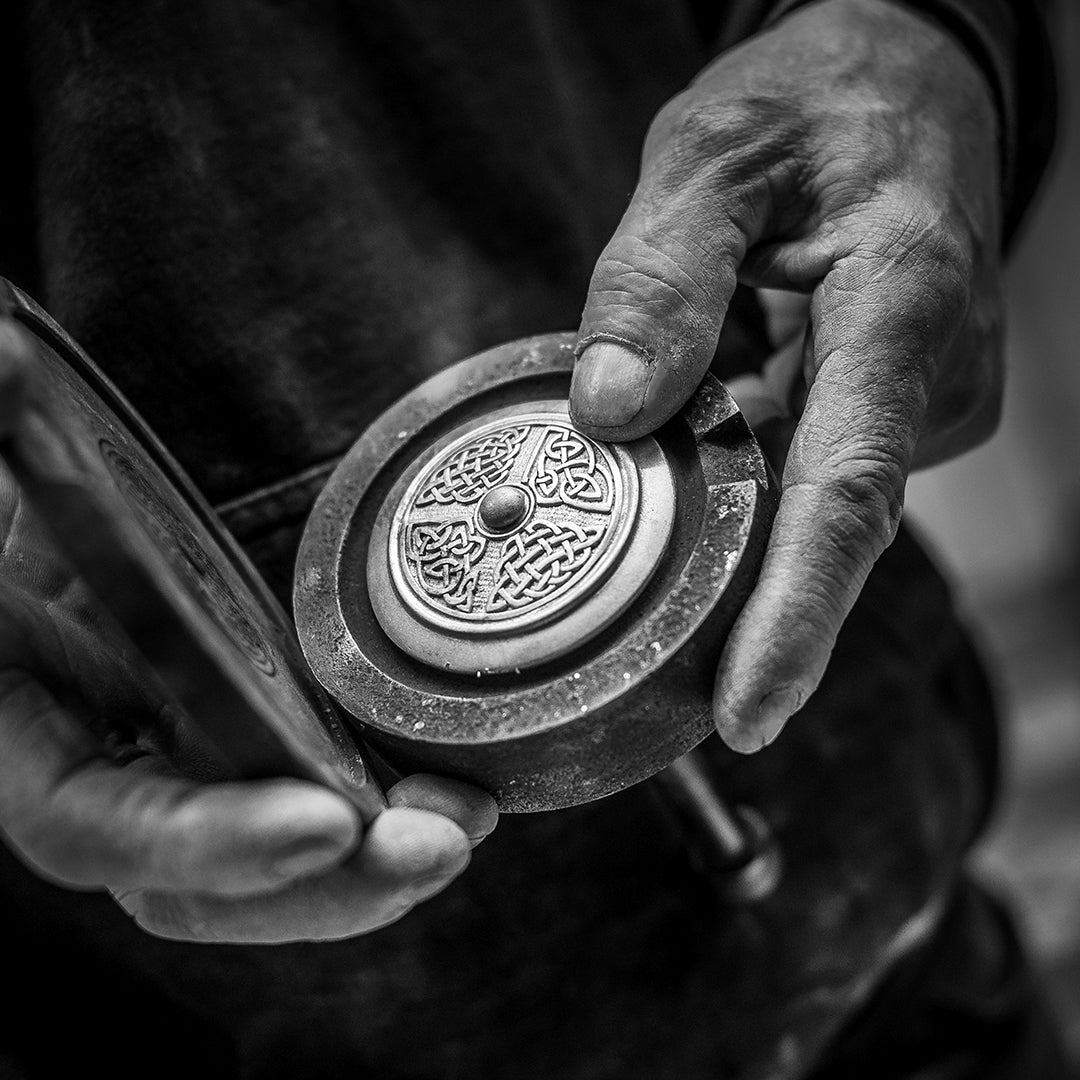 A close-up black and white photograph of a person's hands holding a round metal object with an ornate engraved design on its face.