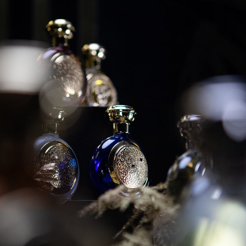 A close-up, shallow depth-of-field shot of several ornate perfume bottles with metallic gold caps and circular silver-toned labels, with one prominent deep blue bottle in sharp focus against a dark background.
