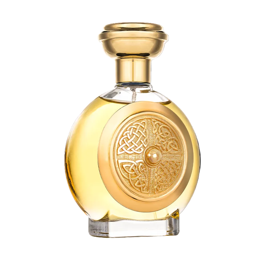 A round glass perfume bottle filled with golden-yellow liquid, featuring a polished gold-toned cap and a circular embossed Celtic knot design on the front, isolated on a white background.