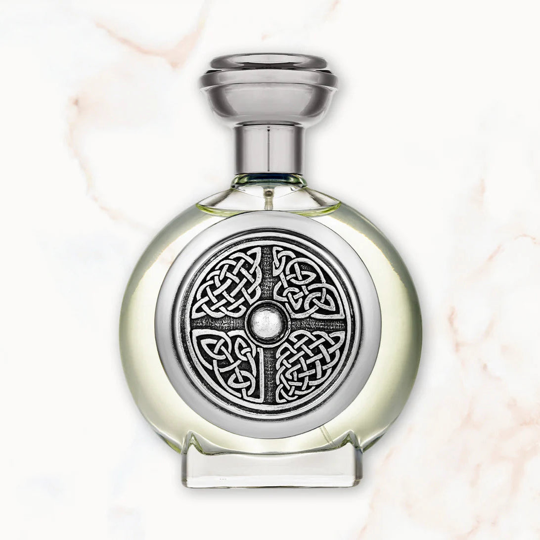 A round glass bottle with a silver-toned metal cap and a decorative silver Celtic knot medallion on the front, shown on a light marble-patterned background.
