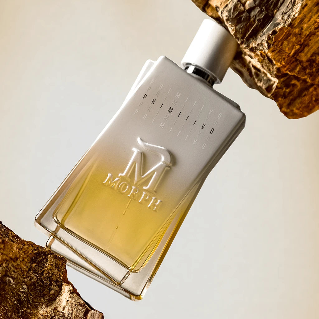 A bottle of Möth Primilius Eau de Toilette perfume with a silver cap and a clear glass body showing a pale yellow liquid, styled resting against textured brown bark on a light beige background.