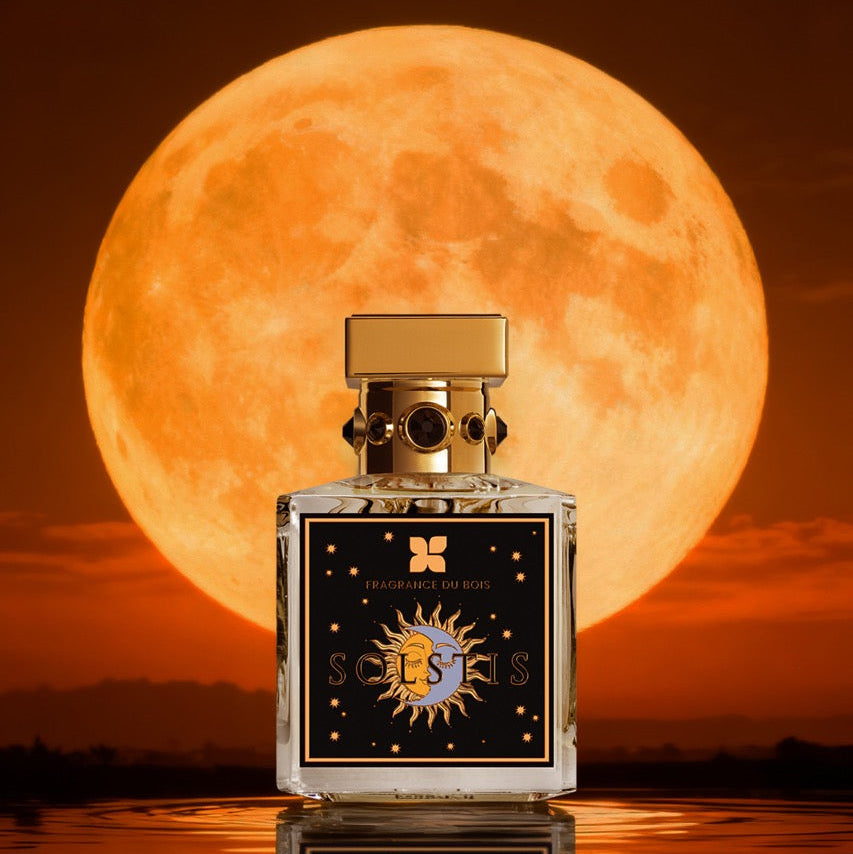A square glass perfume bottle with a gold cap and a black decorative label featuring a gold sun emblem, set against a giant glowing orange full moon over reflective water.