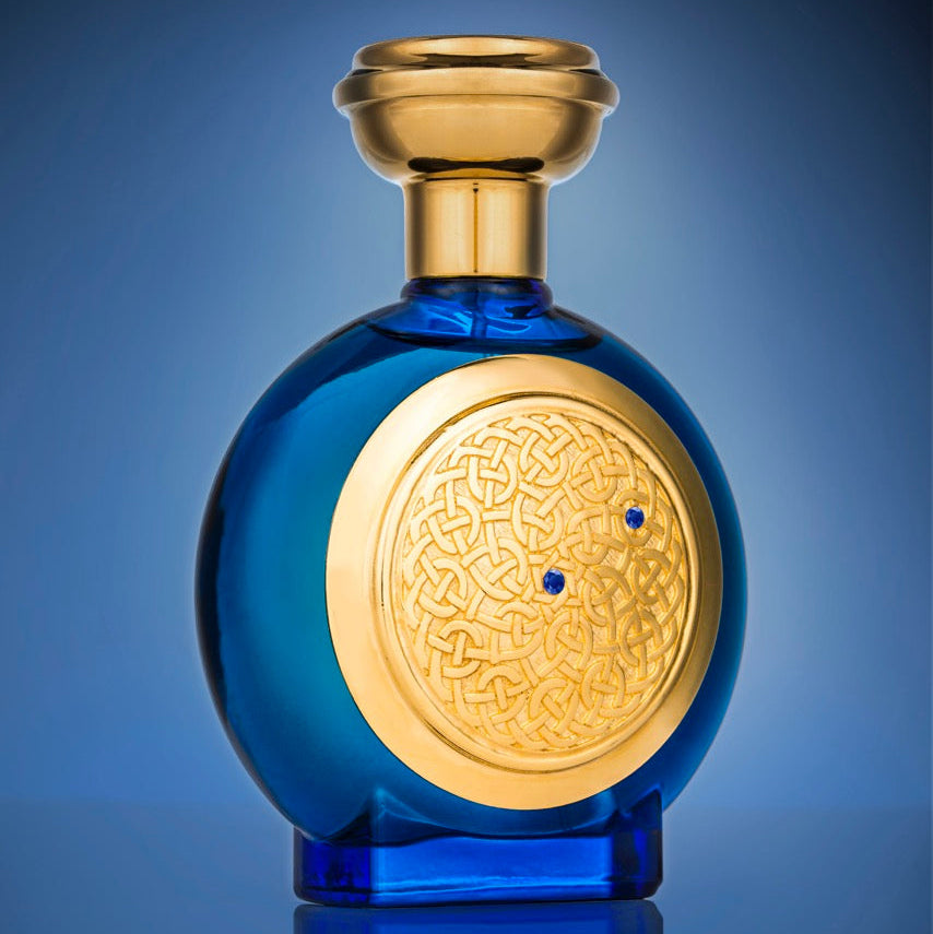 A round blue glass perfume bottle with a gold cap and a large gold circular medallion on the front featuring intricate Arabic calligraphy and two small blue dots, set against a blue gradient background with a reflection below.