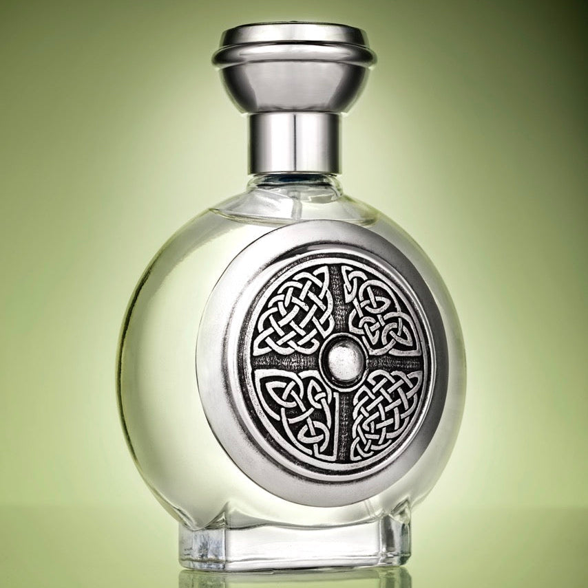 A clear glass bottle with a rounded body, a silver-toned cap, and a large circular Celtic knot design on the front, shown against a green gradient background with a reflection below.