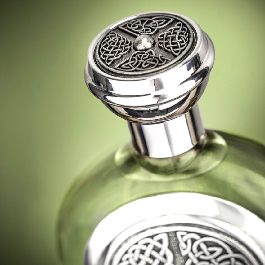 A close-up of a green glass bottle with a highly polished silver-toned metal cap and matching silver-toned circular emblem on the neck, set against a soft green gradient background.